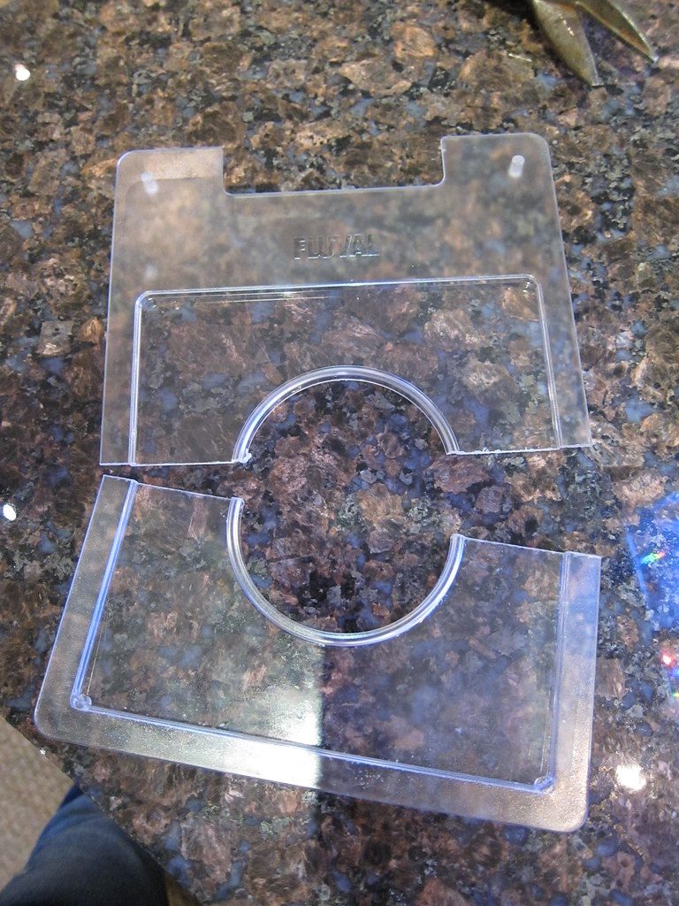 A clear plastic aquarium lid lying on a stone counter, showing a large circular cutout in the middle and two rectangular notches along one edge.
